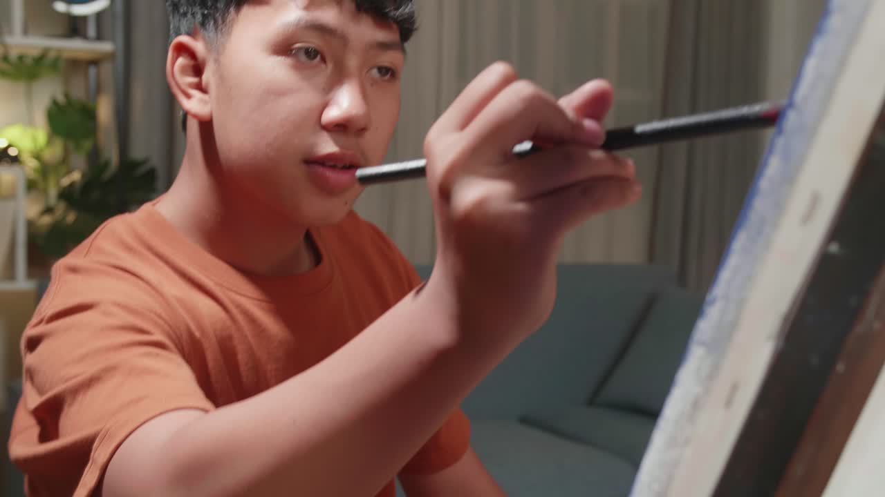 Asian Artist Boy Sitting On Round Wooden Chair Without Backrest Holding Paintbrush And Mixing It With Colour Before Painting On The Canvas