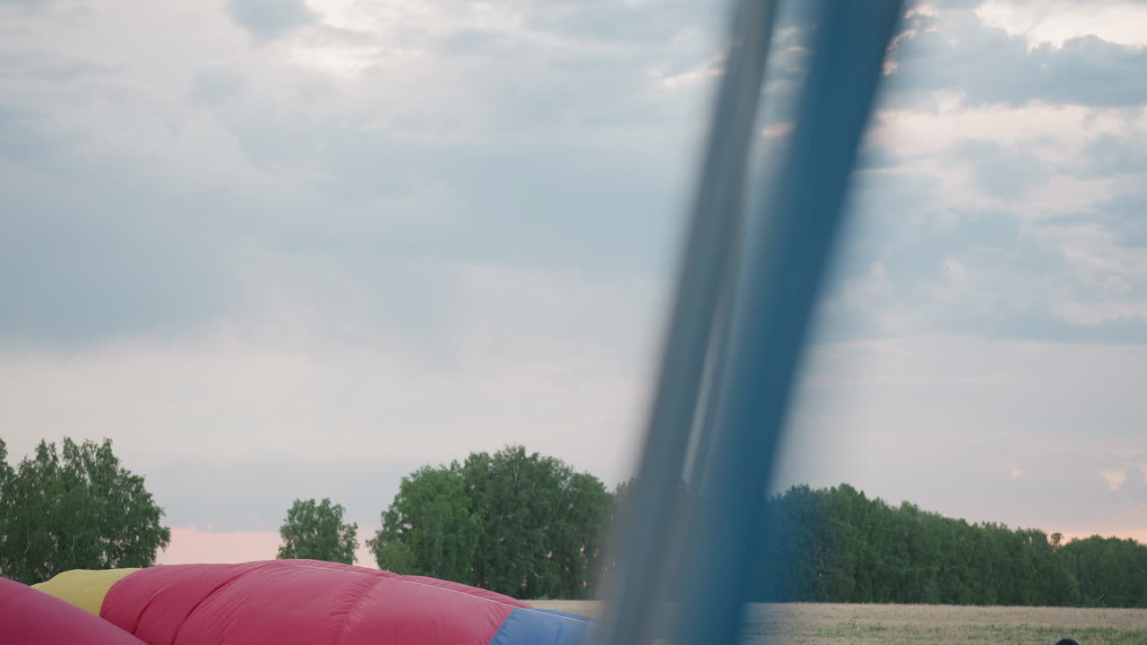 distant tree line silhouettes framing pastel evening sky over open grassy field with partial view of colorful balloon envelope edge and burner frame poles in foreground