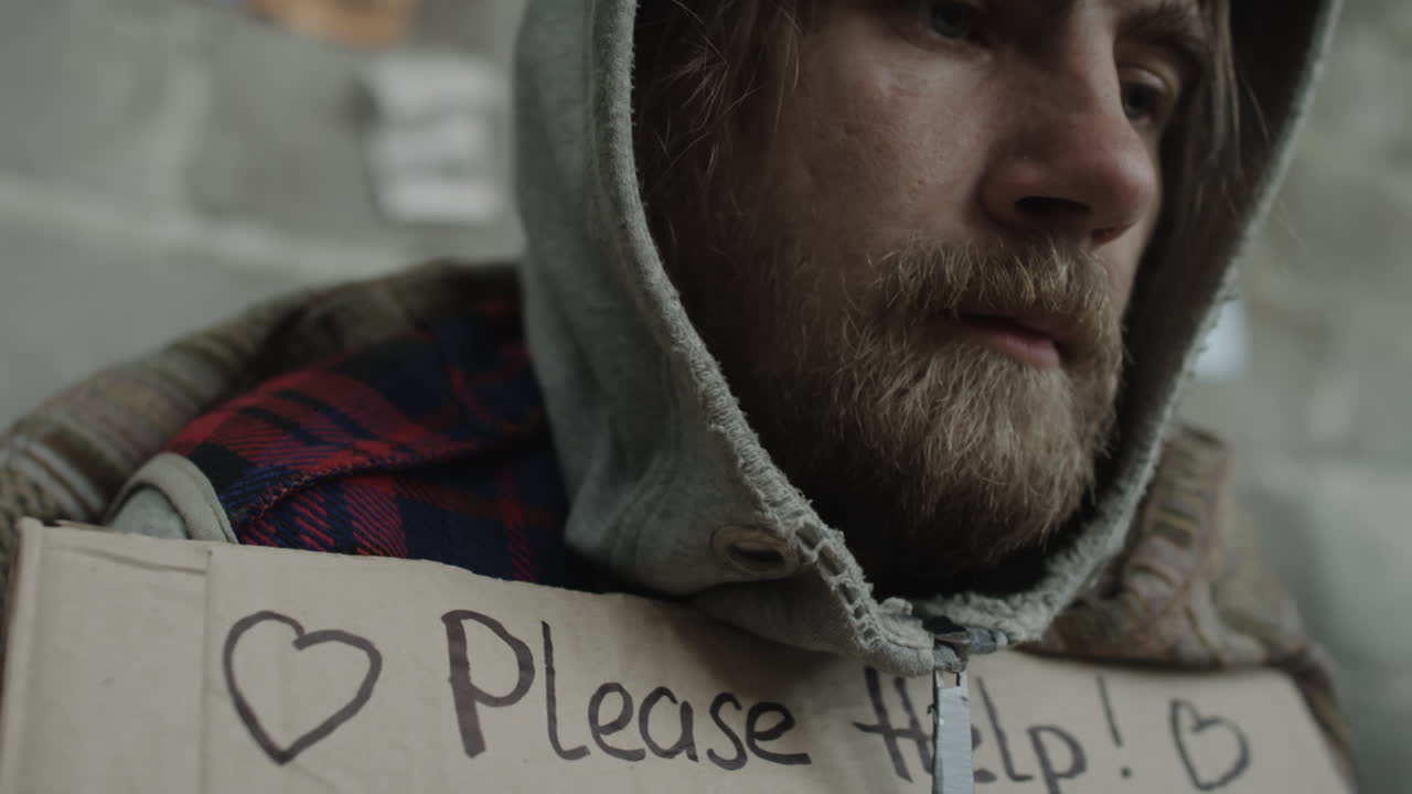 Close-up of a man holding a 'Please Help! Homeless Hungry' sign looking pensive