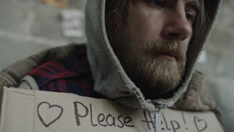 close-up of a man holding a 'please help! homeless hungry' sign looking pensive