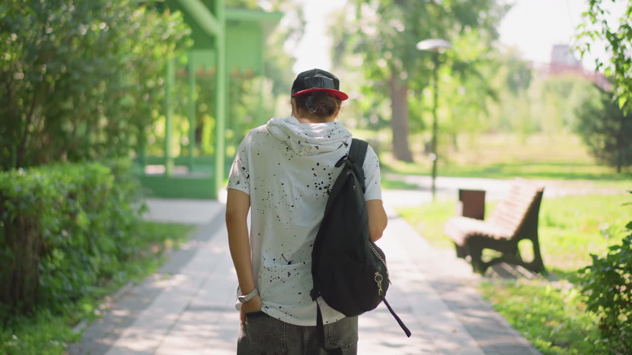 젊은 남자가 나무가 늘어선 길을 걸어가고, 여행자는 백팩을 정리하고, 구경하는 사람은 발걸음을 멈춰 올려다보고, 얼룩덜룩한 빛 속에서 사색에 잠긴 분위기, 조용한 벤치와 멀리 보이는 스카이라인, 느리고 사색적인 발걸음