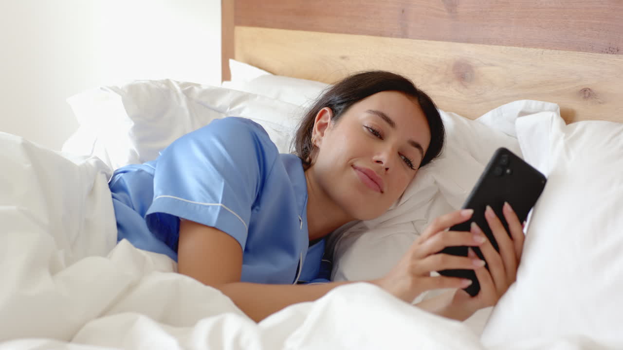 Woman in blue pajamas laughing while using smartphone in bed at home
