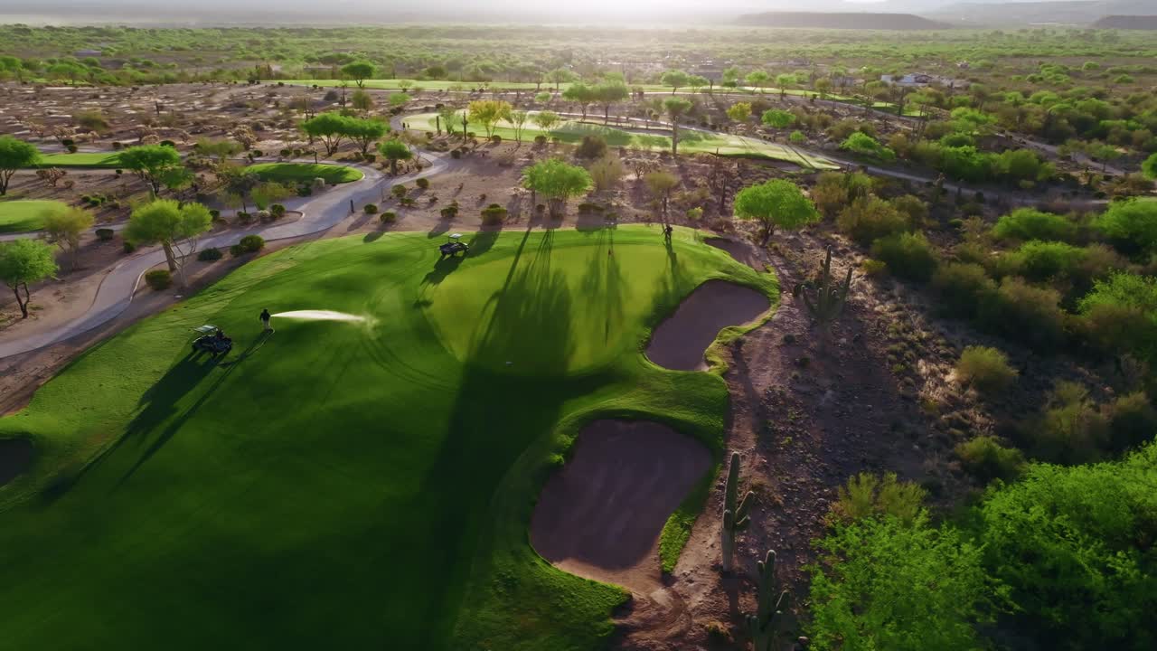 Orbiting drone shot of a desert golf course with bunker, flagstick, green fairway, and cacti backed by rugged mountains as greenskeepers tend and water the green