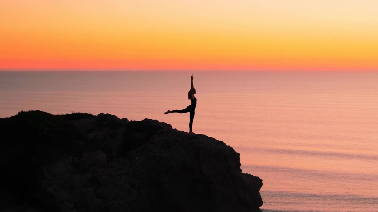 Al centro della clip una persona solitaria si bilancia in una posa yoga sul bordo di una scogliera frastagliata sopra un mare tranquillo