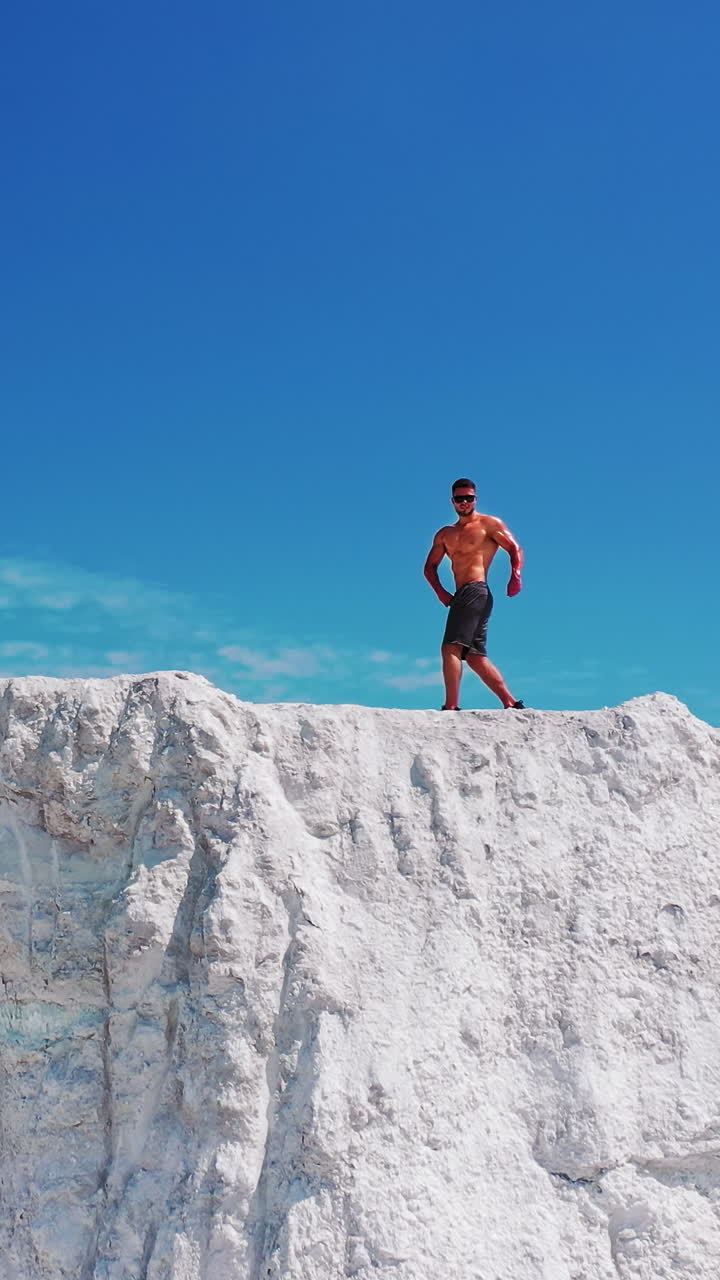 Muscular sportsman under blue sky. Strong athlete in black sunglasses posing on camera on the top of white mountain. Camera moves forward. Drone view. Vertical video