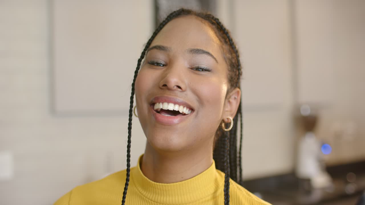 Femme commençant à parler dans la cuisine, souriant, montrant des cheveux tressés et des boucles d’oreilles près de la cafetière