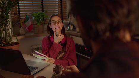 Over-the-shoulder-a-confident-brunette-girl-in-glasses-and-red-wireless-headphones-in-a-sweater-studies-foreign-languages-with-her-teacher-and-learns-the-correct-pronunciation-with-the-help-of-special-exercises-in-a-modern-apartment-in-the-kitchen-near-the-flag-of-Italy