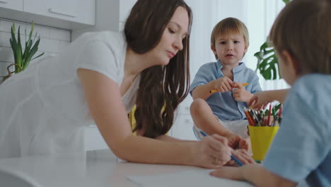 mamá ayuda a los hijos a aprender a dibujar haciendo la tarea preescolar preparación en casa sentados en la cocina blanca. dos niños hermanos dibujan un retrato de la madre juntos