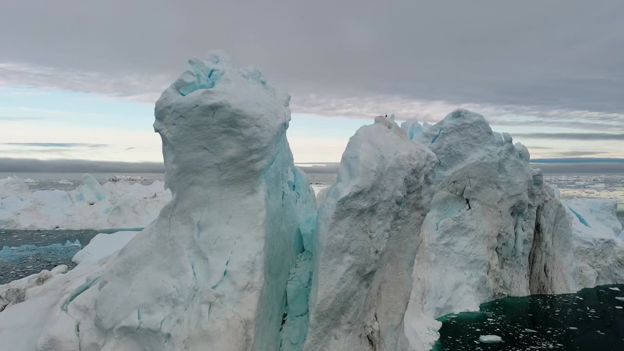 Aerial drone view of a massive towering iceberg floating in the calm Arctic sea near Greenland, showing striking textures, blue ice details, and the frozen beauty of polar nature