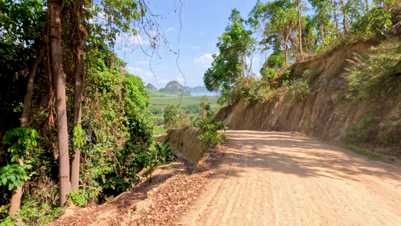 Sunny daytime camera glides along rural dirt road, revealing tropical jungle and distant limestone mountains