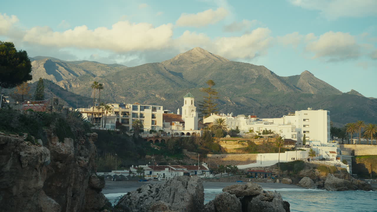 Blick auf die Touristenstadt Nerja zur goldenen Stunde mit weiß getünchten Gebäuden, Kirchturm, felsigen Klippen und dem Mittelmeer, umrahmt von dramatischen andalusischen Bergen