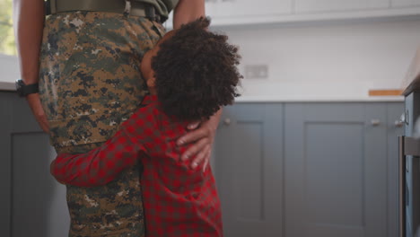 close up of son hugging legs of army mother in uniform home on leave in family kitchen