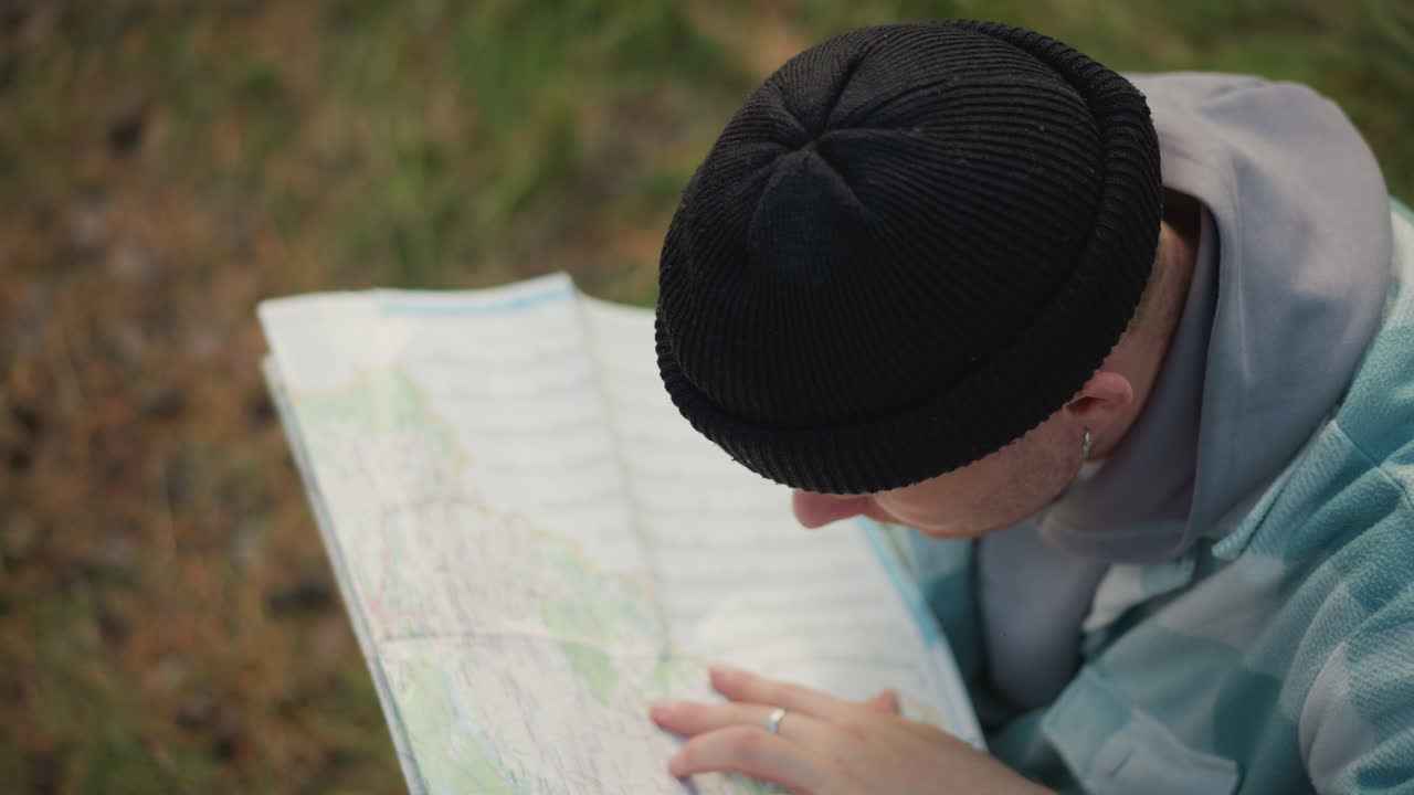 white male explorer confirming map route by finger trace on paper, decisive moment in forest clearing, tattooed hand points to waypoint, wearing beanie and plaid jacket with backpack nearby, closeup