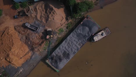 aerial view loading gravel by crane from a river barge to truck establishing shot with tug boat and interesting shadows