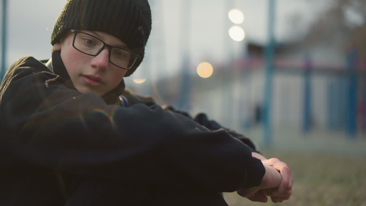 Close-up of a young boy wearing glasses and a black beanie with grass particles on his head, sitting and speaking with a blurred background