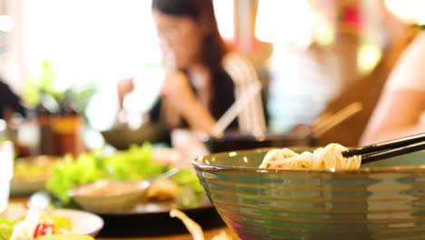 chopsticks lift noodles from a bowl in a bustling vietnamese restaurant, capturing the vibrant dining atmosphere with bright lighting and lively ambiance