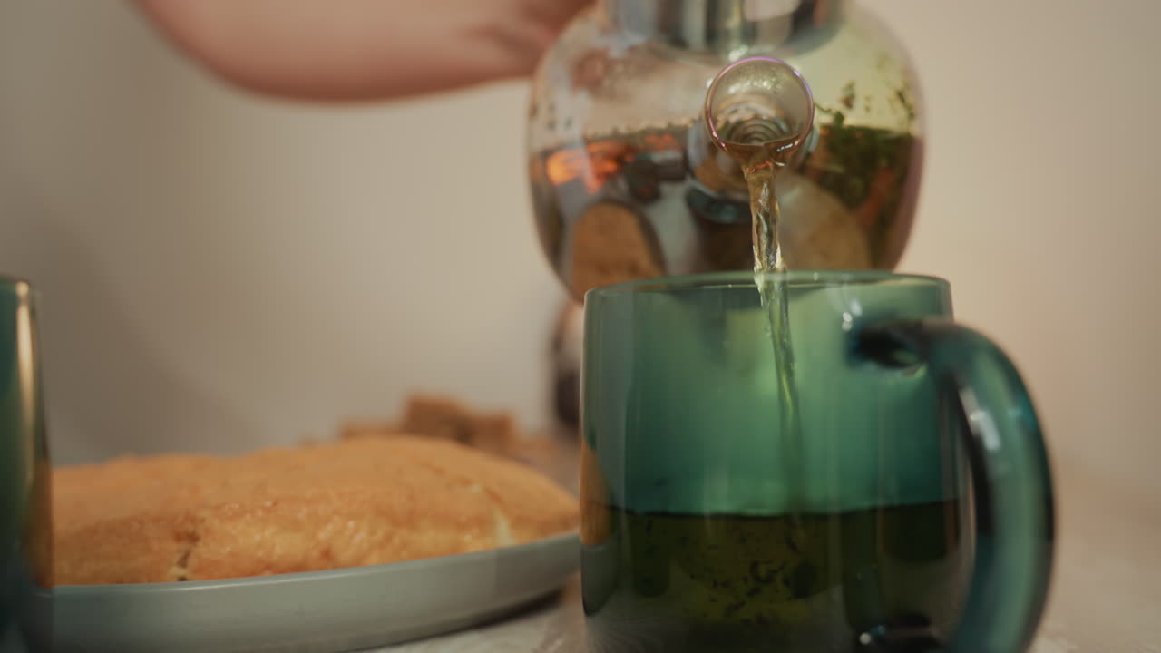 tazza verde che riceve tè alle erbe fumante versato da una mano delicata da una teiera di vetro accanto a una torta appena sfornata su un vassoio di metallo, illuminazione soffusa e calda, bassa profondità di campo, tavolo da cucina intimo, accogliente