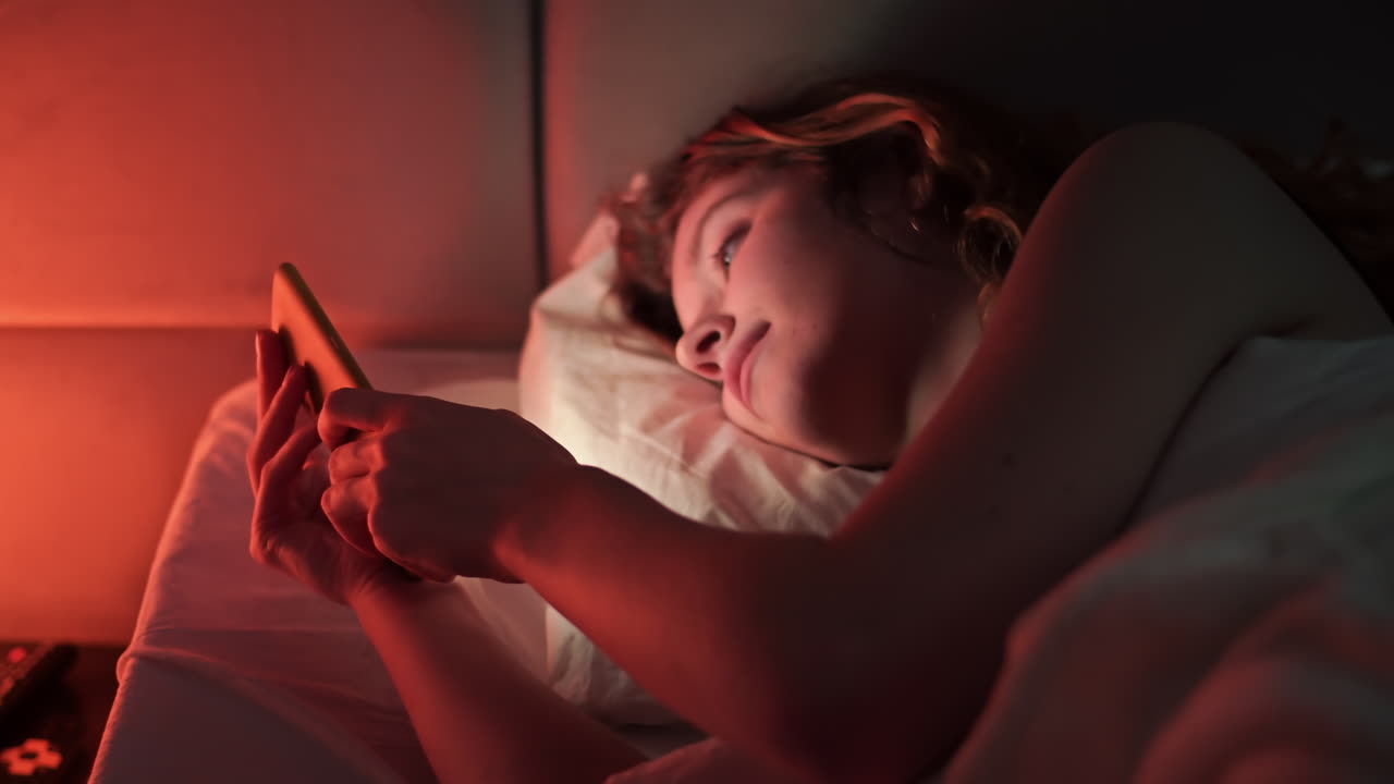 Young ginger hair girl browsing her smartphone at night in the bed. Colorful light