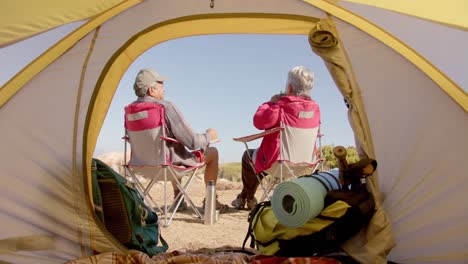 Happy-senior-biracial-couple-sitting-at-tent-in-mountains-and-drinking-coffee,-in-slow-motion