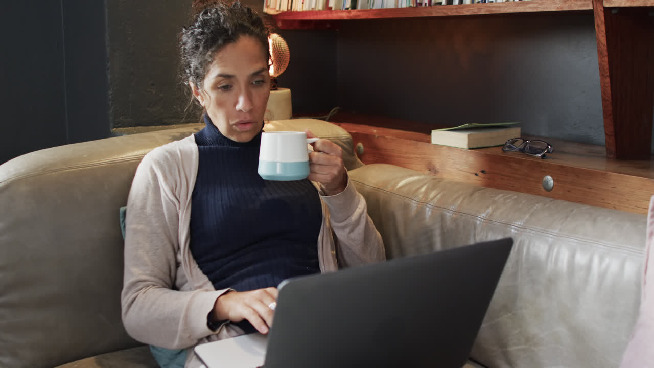 Biracial woman sitting on sofa using laptop and drinking coffee at home, slow motion