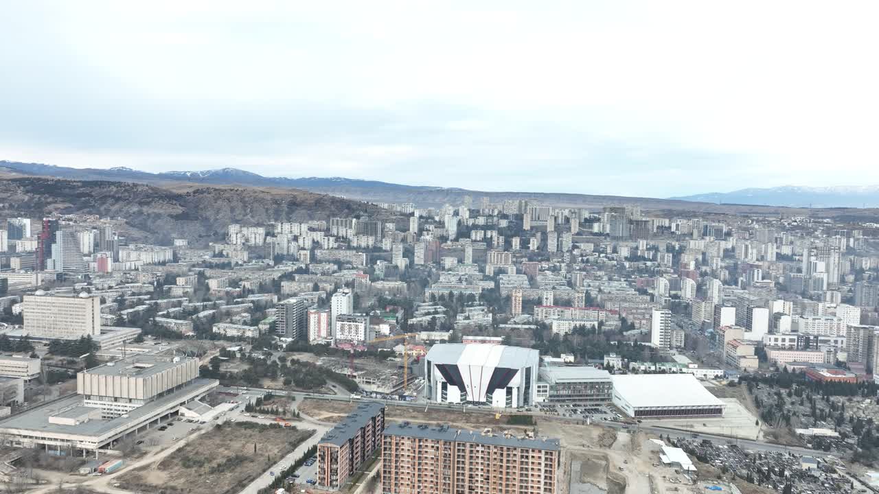 An aerial perspective of a modern cityscape, showcasing a mix of high-rises, residential areas, and industrial zones. The muted winter tones and distant hills frame the urban horizon