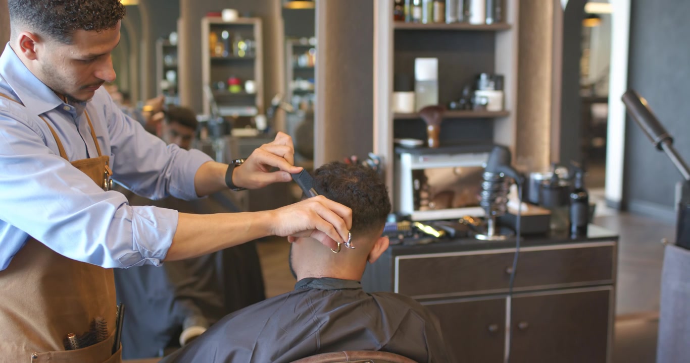 Afroamerikansk barber sætter trimmeren i nakken, klipper og fader en kunde i salonen, iført forklæde