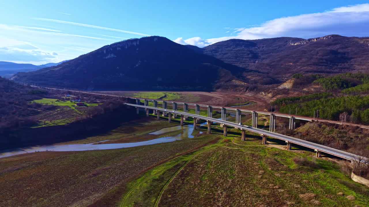 Vista di un ponte che collega due colline sopra un fiume e un terreno durante una giornata serena in una zona rurale con montagne in lontananza