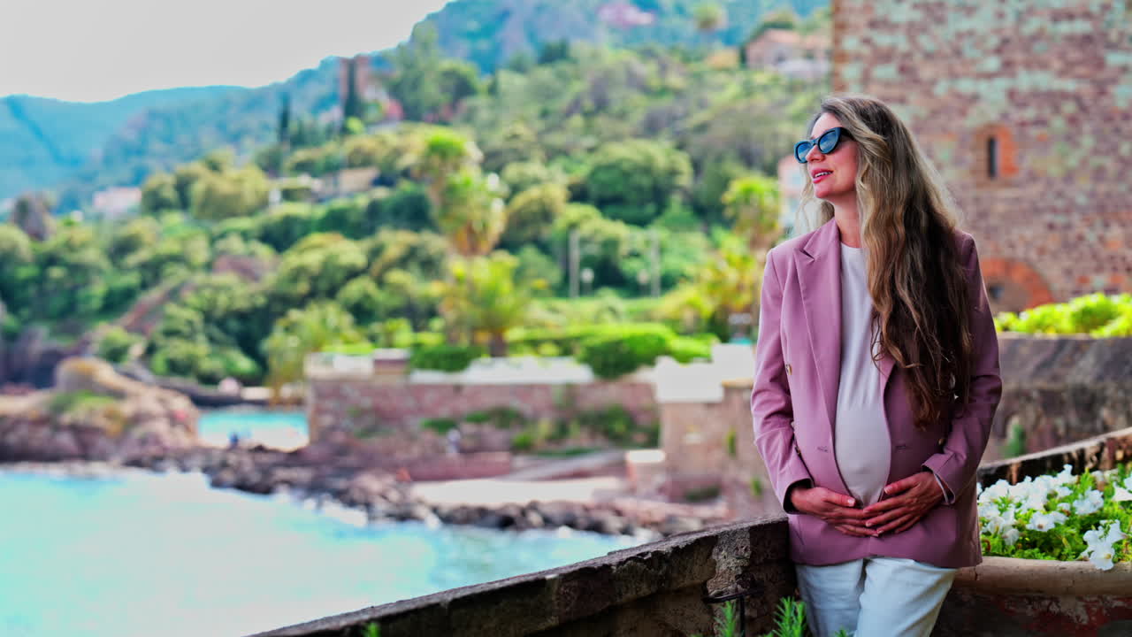 Pregnant woman with long hair, wearing sunglasses standing with the Chateau de la Napoule Castle in Mandelieu-La Napoule, France in the background