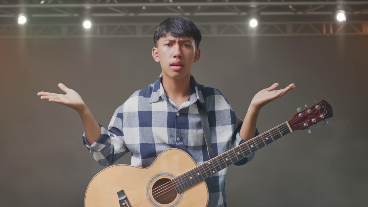 기타를 들고 있는 아시아인 십대는 당황스러운 몸짓을 하고 무대에서 뭐라고 말하고 있습니다.