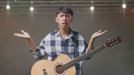 adolescente asiático con guitarra mostrando un gesto desconcertado y diciendo qué en el escenario