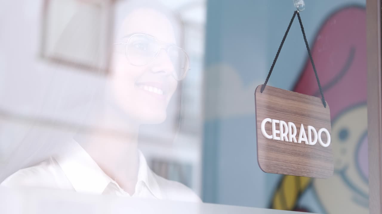 Happy small business girl changing closed to open sign in spanish, smiling looking outside waiting for clients. Start-up concept. 4k 60fps