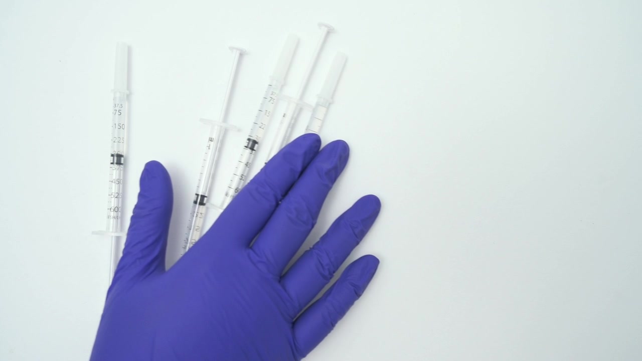 Mano di un operatore sanitario con un guanto sterile che prende siringhe da una superficie su sfondo bianco.