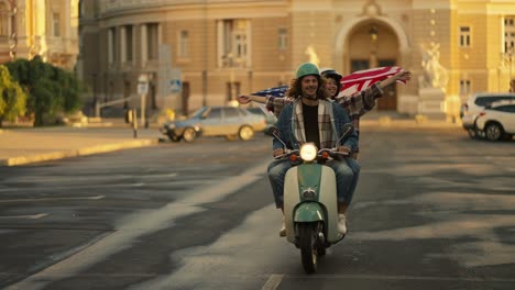 A-brunette-guy-with-curly-long-hair-in-a-denim-jacket-rides-with-his-girlfriend-who-is-holding-a-large-US-flag-in-her-hands-and-it-flutters-in-the-wind-during-their-ride-in-the-summer-city