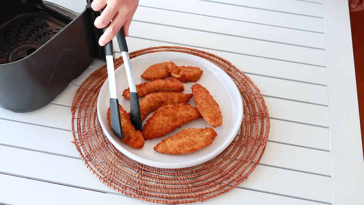 Golden chicken tenders placed on a plate using tongs, next to an air fryer on a white table