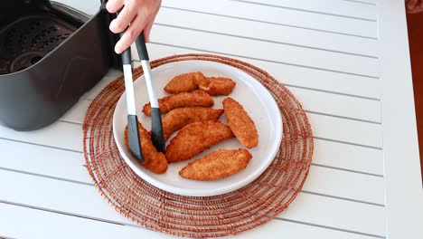 golden chicken tenders placed on a plate using tongs, next to an air fryer on a white table