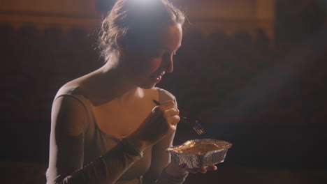 beautiful ballerina eats and discusses performance with partner after choreography rehearsal. ballet female dancer sits on theatre stage. empty rows in theatrical hall. ballet concept. portrait.