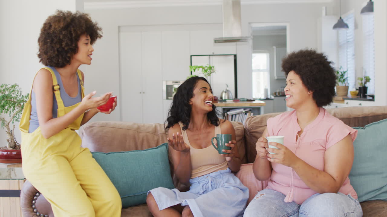 felices amigas diversas sentadas en el sofá en la sala de estar, hablando y sonriendo