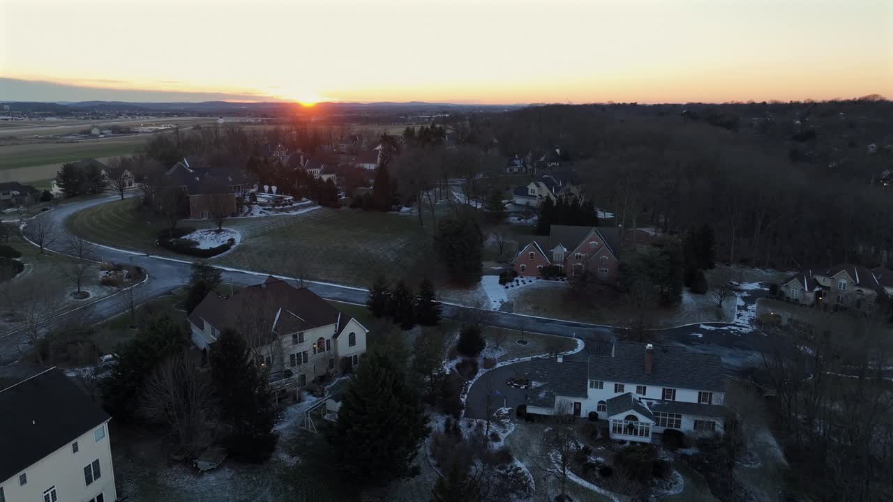 Luftfoto af det landlige Amerika ved solnedgang om vinteren, med sneklædte palæer og huse omgivet af træer og åbne marker, mens et varmt lys forsvinder over et stille landskab. Bredt billede
