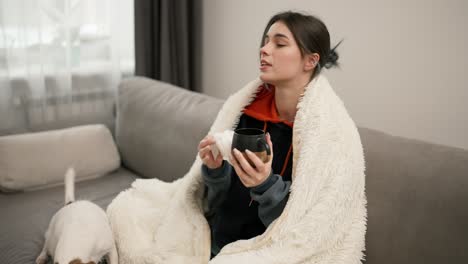 Sick-pretty-girl-suffering-with-flu,-drinking-hot-tea-and-warming-up-with-blanket-on-sofa-with-terrier-dog