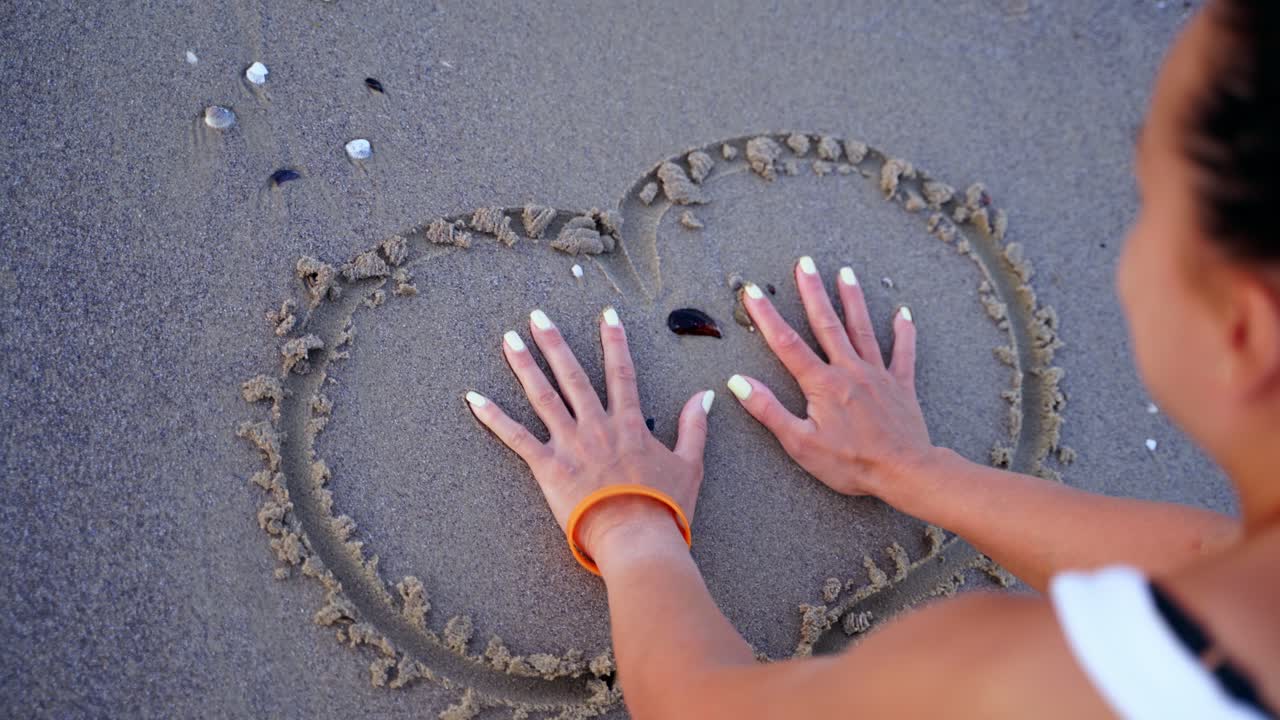 Woman is painting on wet sand. Top view of a heart written on a sandy beach. Young female drawing near the sea.