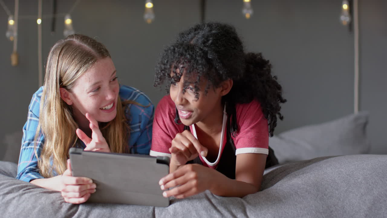 Happy diverse female friends lying on bed and using tablet at home, slow motion