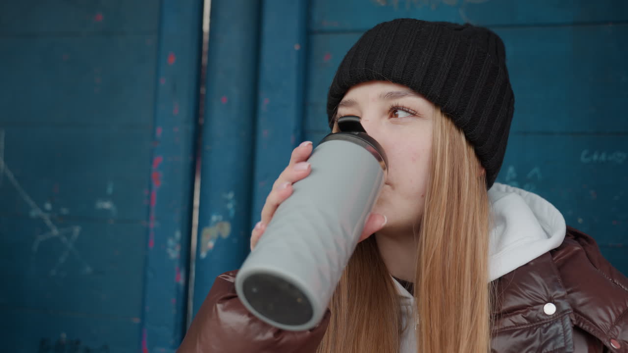 갈색 코트와 검은색 비니를 착용한 백인 여성이 파란색 벽 옆, 조용한 야외 겨울 쉼터에서 여행용 머그에 담긴 따뜻한 음료를 마시고 있어요. 긴 머리가 보이고, 통근 중 휴식을 취하며 편안한 표정을 짓고 있습니다.