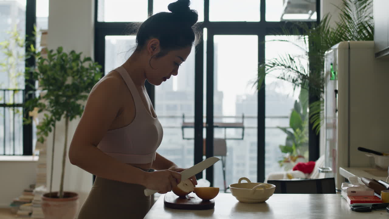 donna asiatica al rallentatore che taglia frutta di pera nella sua moderna cucina in un appartamento con paesaggio urbano della città moderna vista dalla finestra