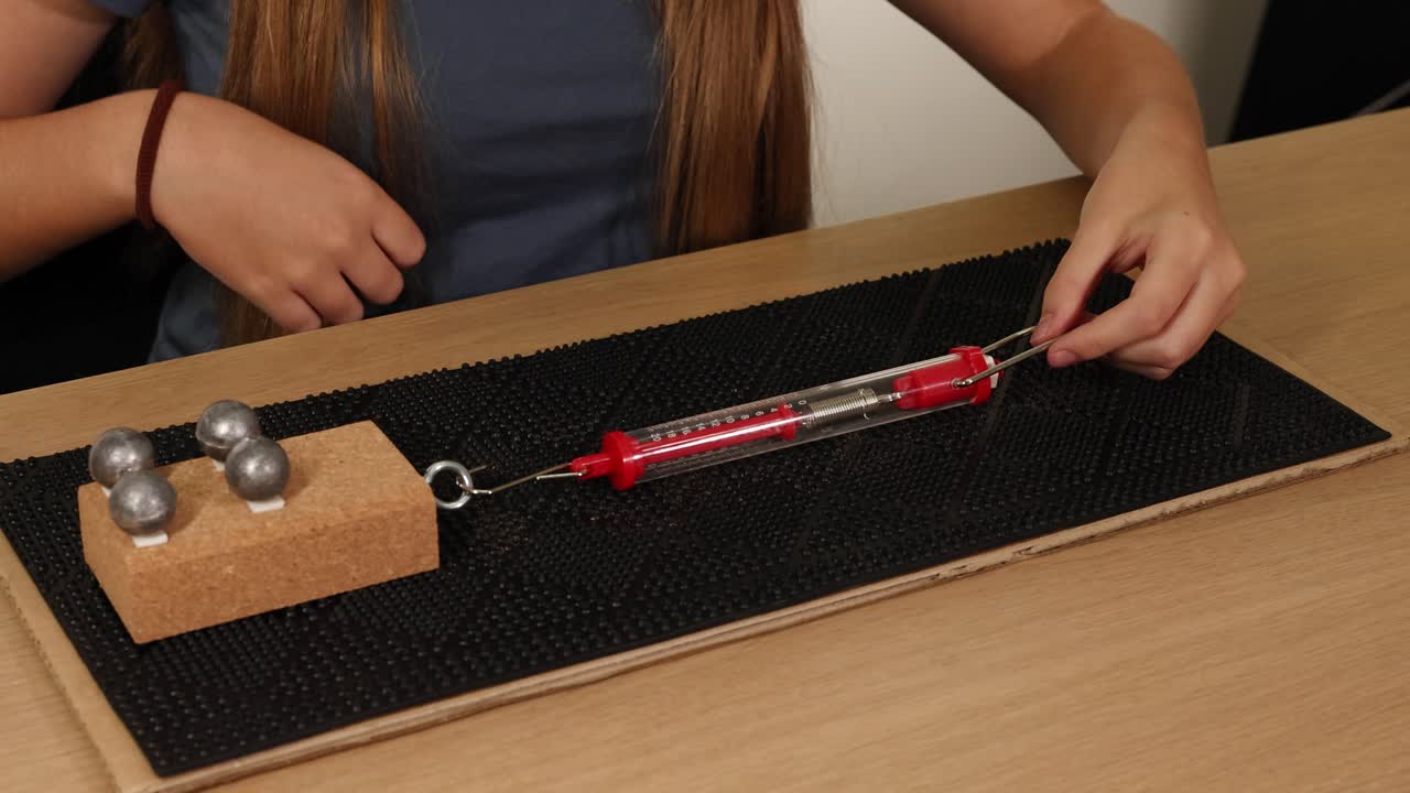 Hands use a spring scale to measure force needed to drag weights on a cardboard surface, illustrating friction principles