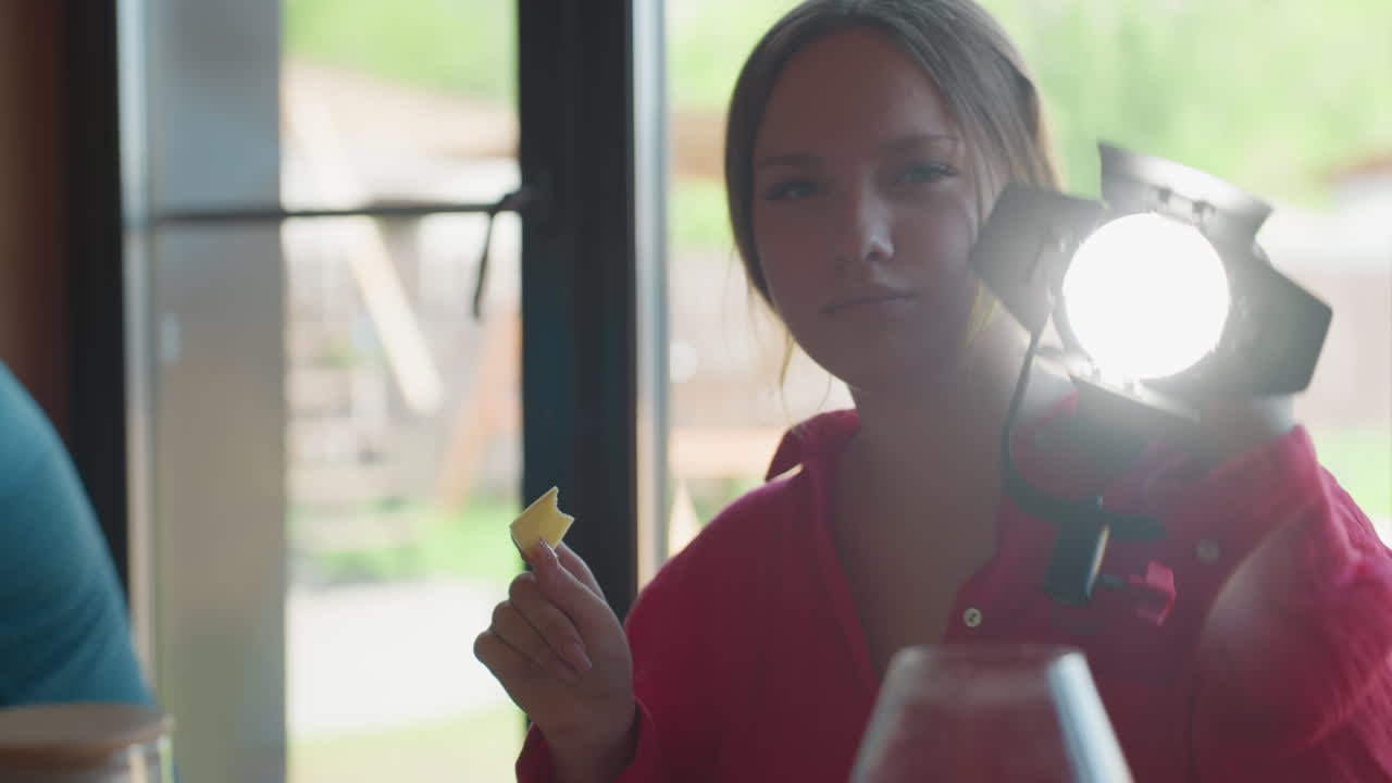 Young lady in red shirt smiles while eating sliced cheese and holding studio spotlight on shoulder, standing near large window, soft natural light blends with beam