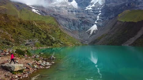 aéreo, disparo de un dron siguiendo a una mujer caminando en la orilla rocosa del lago humantay, día nublado, en los andes, región cusco, perú