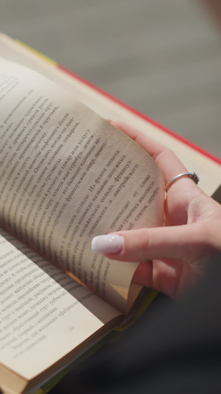 primer plano de las manos de una mujer girando una página de su libro al aire libre, uñas pintadas con esmalte blanco, libro de bordes amarillos abierto, mostrando texto en las páginas
