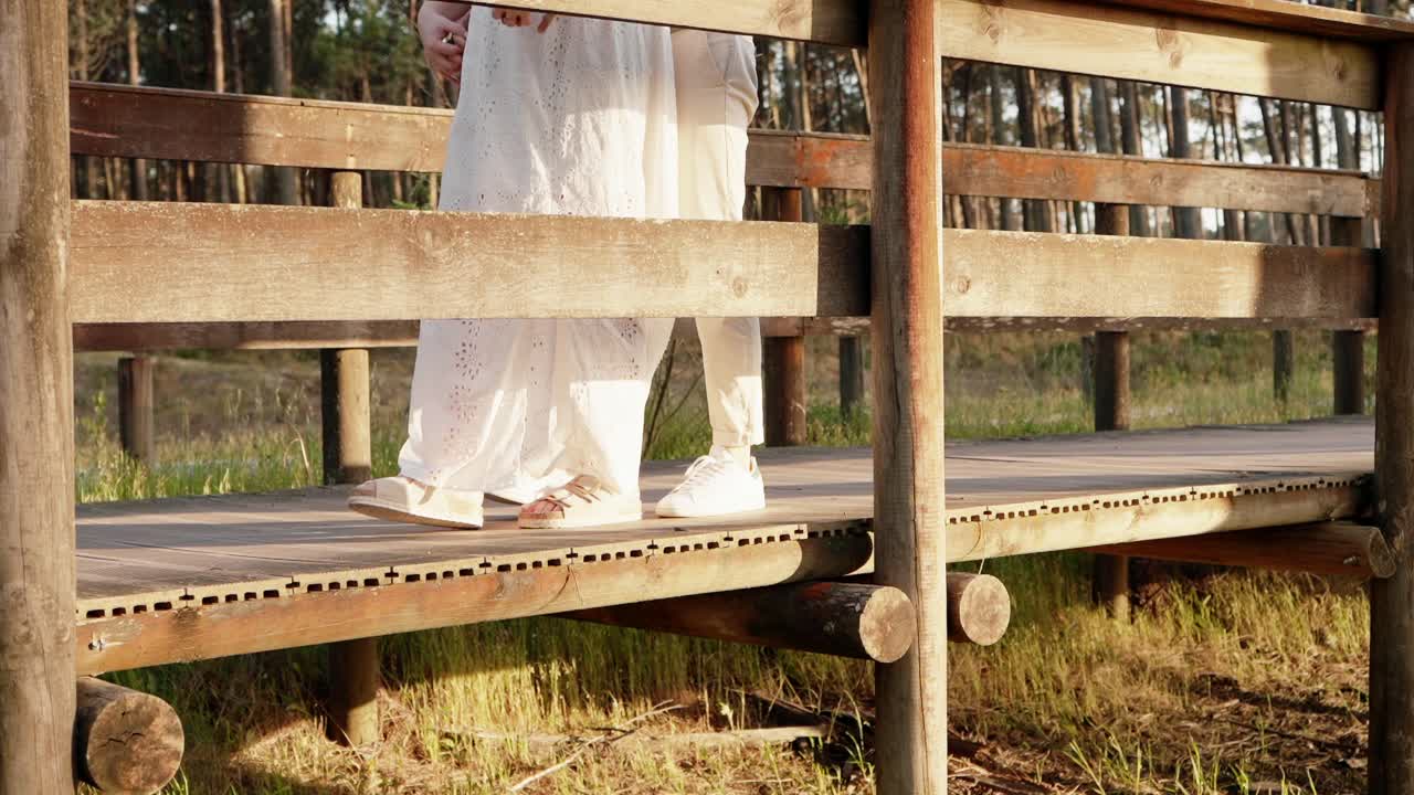 Bride in white dress and sneakers walking on a wooden bridge through grassy forest path
