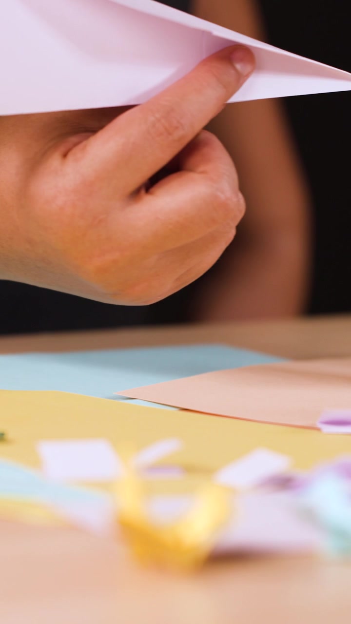 Primo piano di mani che piegano carta bianca per creare un aeroplanino di carta origami su un tavolo di legno.