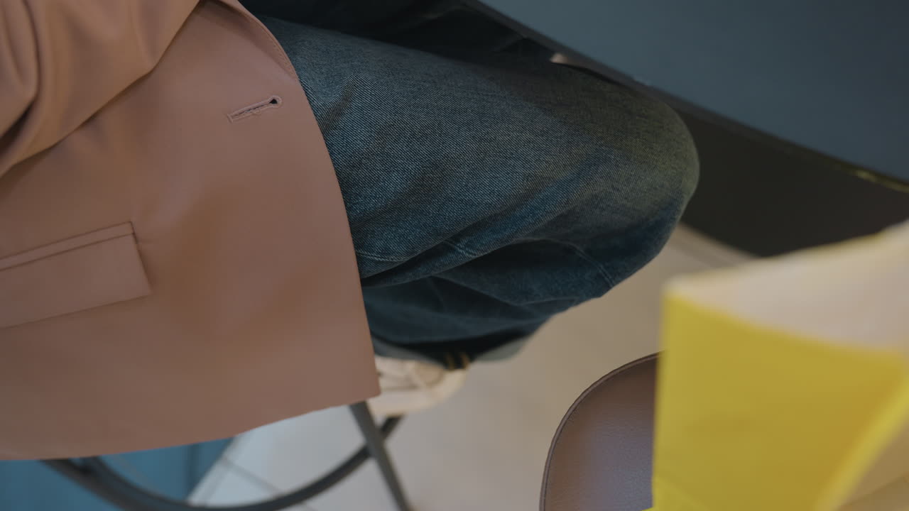 Persona sentada con accesorios en una cafetería, persona descansando con un bolso y una americana en un ambiente de cafetería moderna, persona informal con vaqueros y americana esperando pacientemente en una mesa de cafetería contemporánea